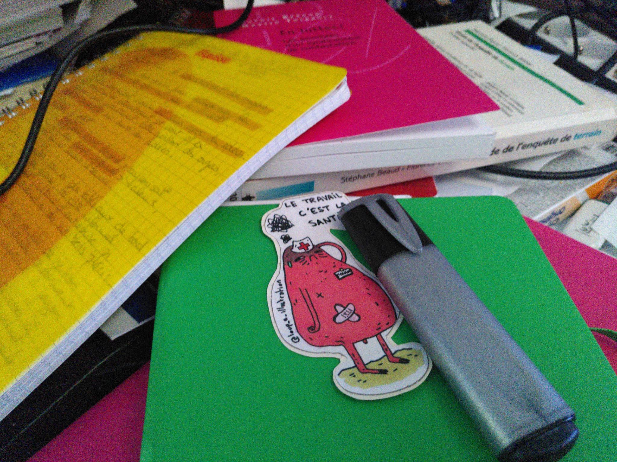 Mon bureau en bazar, avec des papiers imprimés, mon agenda, un autocollant “le travail c’est la santé”, un surligneur…